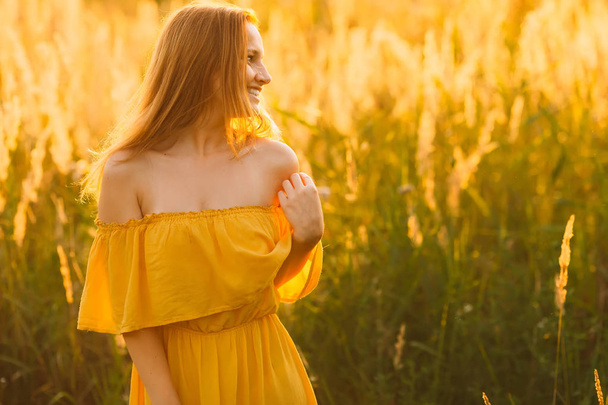 милая девушка в сексуальном платье с открытым плечом повернула голову в сторону и смеется на фоне высокой травы
 - Фото, изображение