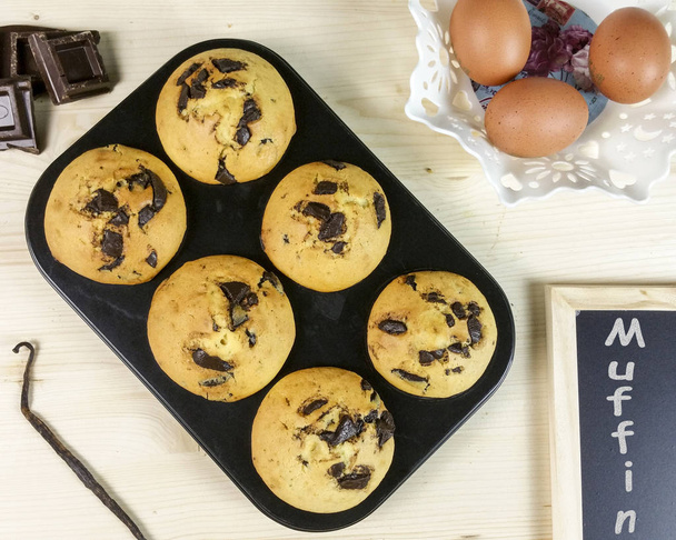 Muffin mit Schokoladentropfen in einer Muffinpfanne, mit Eiern und Vanilleschote auf Holzhintergrund - Draufsicht - Foto, Bild