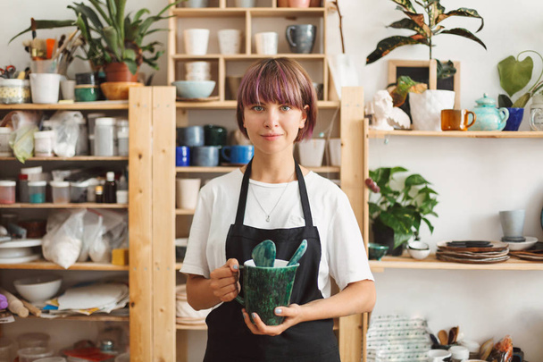 Pretty girl with colorful hair in black apron and white T-shirt holding handmade bowl in hands dreamily looking in camera spending time at modern pottery studio - Foto, afbeelding