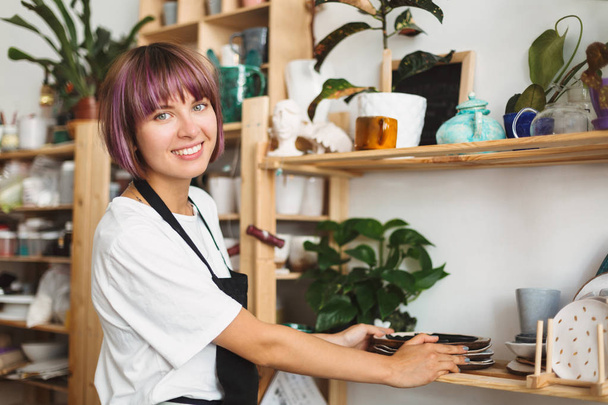 Beautiful smiling girl with colorful hair in black apron and white T-shirt putting handmade plates on shelf joyfully looking in camera at pottery studio - Photo, Image