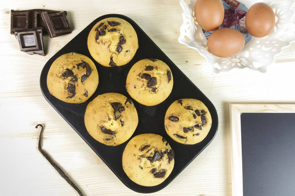 Muffin com gotas de chocolate em uma panela de muffin, com ovos e feijão de baunilha no fundo de madeira vista superior
 - Foto, Imagem