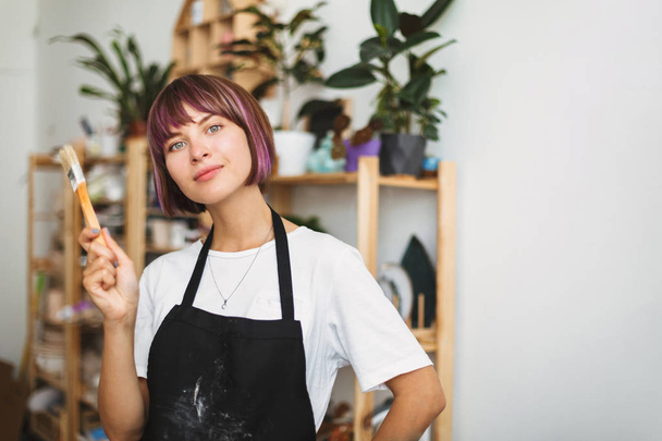 Beautiful girl with colorful hair in black apron and white T-shirt holding paint brush in hand dreamily looking in camera at modern pottery studio - Photo, Image