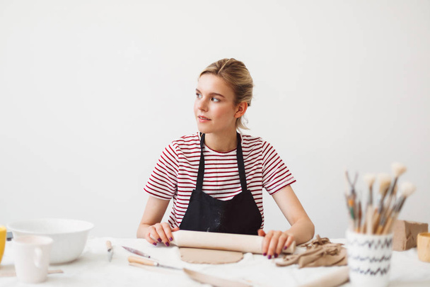 Beautiful girl in black apron and striped T-shirt sitting at the table holding rolling pin working with clay and dreamily looking aside at pottery studio - Photo, Image