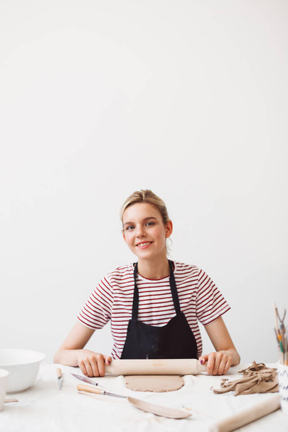 Pretty smiling girl in black apron and striped T-shirt sitting at the table with rolling pin and working with clay joyfully looking in camera at pottery studio - Foto, imagen