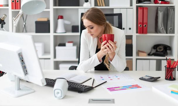 若い女の子は、オフィスのコンピューターのデスクで座って、彼女の手で赤カップを保持します。テーブルの上の少女の前に、図. - 写真・画像