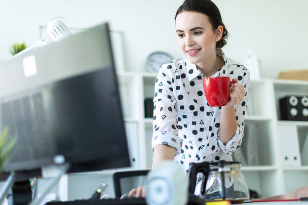 A young girl is standing in the office near the table, holding a red mug in her hand and typing on the keyboard. - Foto, Imagen