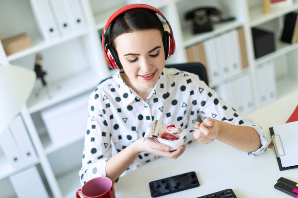 schönes junges Mädchen sitzt mit Kopfhörern am Schreibtisch im Büro und isst Joghurt mit roter Füllung. - Foto, Bild