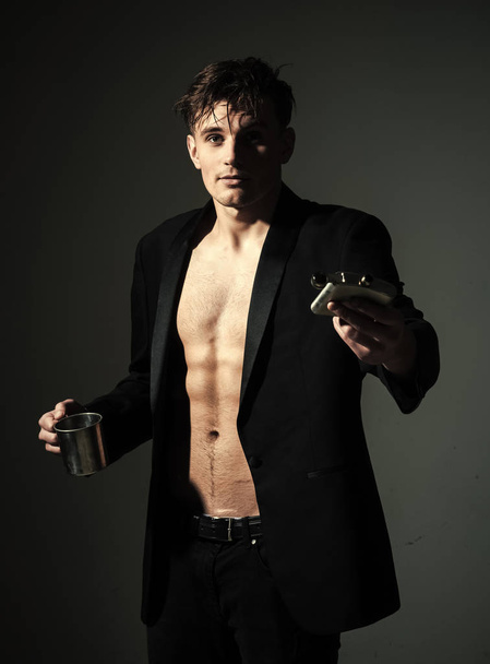 young male alcoholic. man on confident face, muscular figure, holds flask with alcohol and metallic cup. Man with muscular torso, six packs, wears classic jacket, looks attractive, dark background - Photo, image