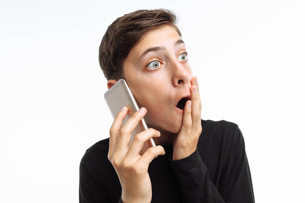 Portrait of a teenager, a guy holding a phone, looking at the phone and shows the emotion of surprise on his face, in a black t-shirt, on a white background, - Фото, изображение