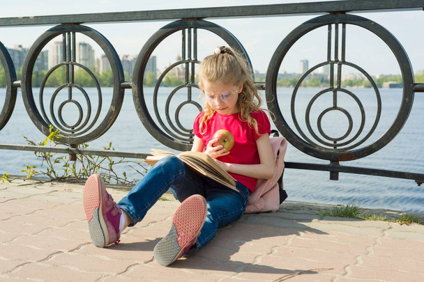 Child girl student wearing glasses with backpack is reading book. Urban background, river, sky. - Fotografie, Obrázek