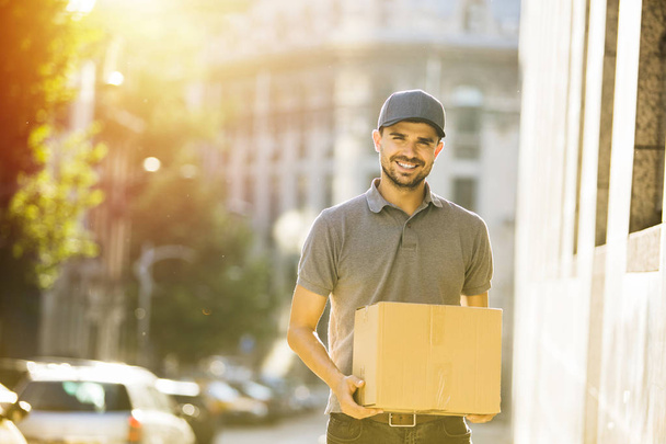 your shipping is here. delivery man in grey shirt with cap going with smile to deliver the package, somewhere on the street - Φωτογραφία, εικόνα