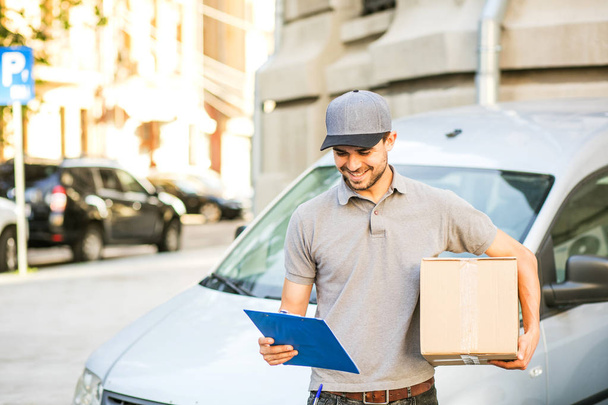 your shipping is here. delivery man in grey shirt with cap standing with his cardboard box looking on the form for last details, in front of his car, outside on the street - Фото, изображение