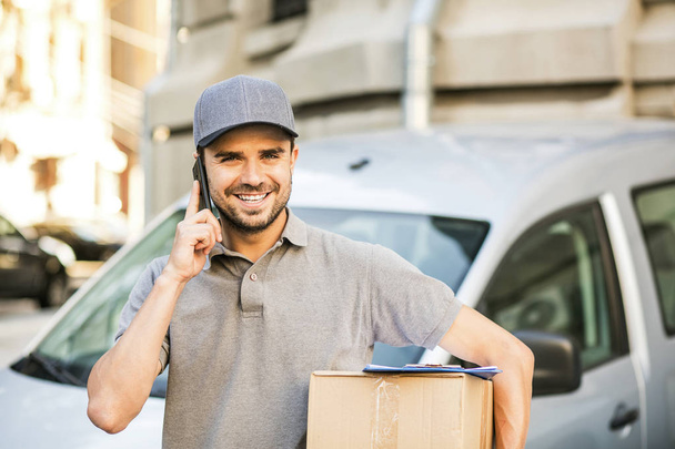 your shipping is here. delivery man in grey shirt with cap standing with his cardboard box on the street calling the client to deliver the package - Photo, Image
