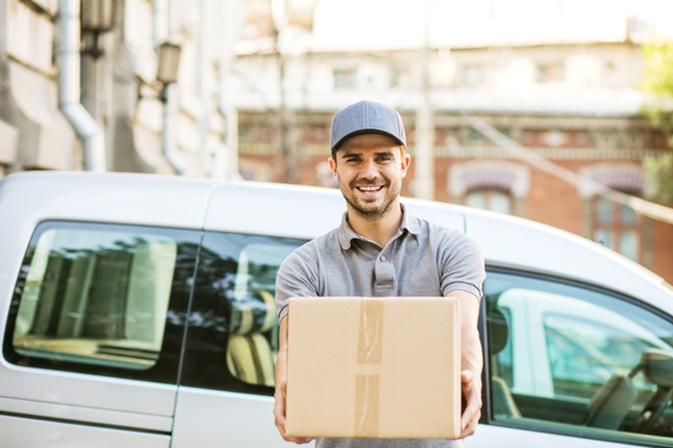 Ваша доставка здесь. счастливый доставщик в серой рубашке и кепке стоит со своей картонкой коробкой на улице, с улыбкой глядя в камеру перед своей машиной доставки
 - Фото, изображение