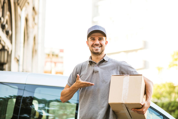 your shipping is here. happy delivery man in grey shirt with cap standing with his cardboard box on the street looking to camera with smile, in front of his delivery car - Fotografie, Obrázek