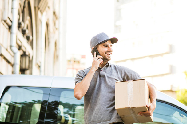 your shipping is here. delivery man in grey shirt with cap standing with his cardboard box on the street calling the client to deliver the package and looking to side - Foto, immagini