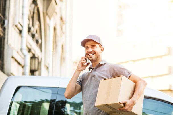 your shipping is here. delivery man in grey shirt with cap standing with his cardboard box on the street calling the client to deliver the package and looking to side - Photo, Image
