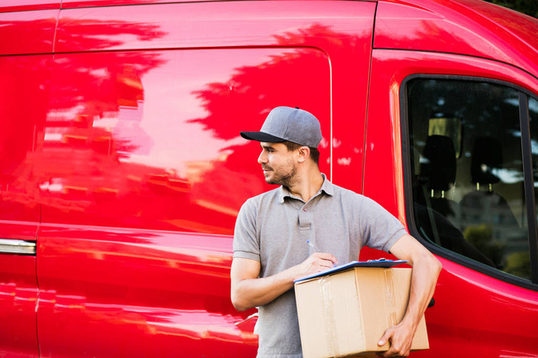 your shipping is here. young delivery man in grey shirt with cap standing with his cardboard box completing the form for deliver, ready to meet the client, outside on the street in front of his car - Photo, Image