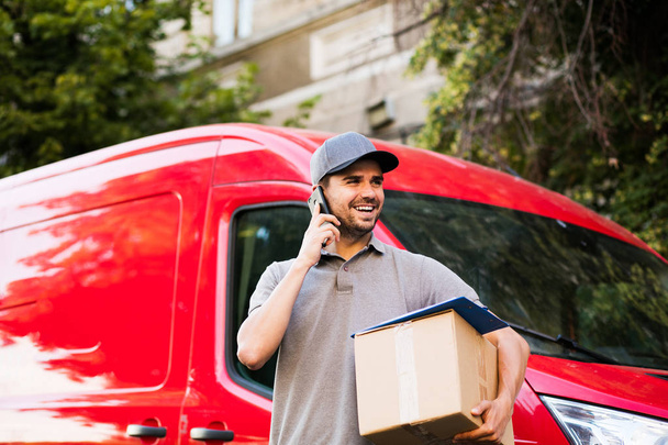 your shipping is here. delivery man in grey shirt with cap standing with his cardboard box on the street calling the client to deliver the package and looking to side - Фото, изображение