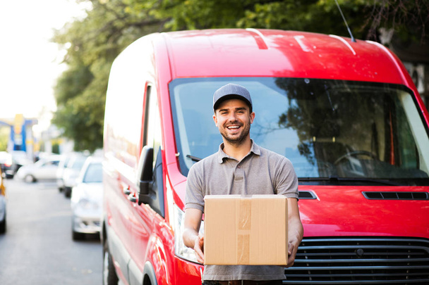 Ваша доставка здесь. счастливый доставщик в серой рубашке и кепке стоит со своей картонкой коробкой на улице, с улыбкой глядя в камеру перед своей машиной доставки
 - Фото, изображение