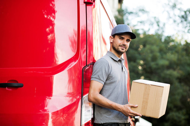 your shipping is here. delivery man in grey shirt with cap standing with his cardboard box at the back of his car, ready to give the package to the client, outside on the street - Foto, imagen