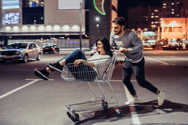 Young romantic couple is having fun with supermarket cart in the evening. Handsome bearded man and attractive young woman are spending time together outdoors. - Fotoğraf, Görsel