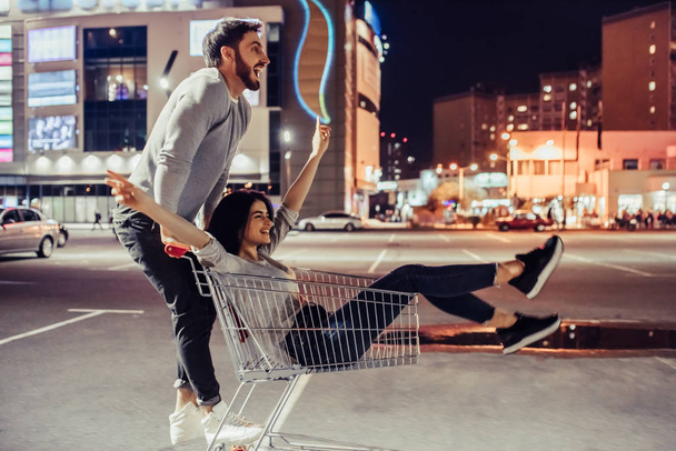 Young romantic couple is having fun with supermarket cart in the evening. Handsome bearded man and attractive young woman are spending time together outdoors. - Foto, Bild