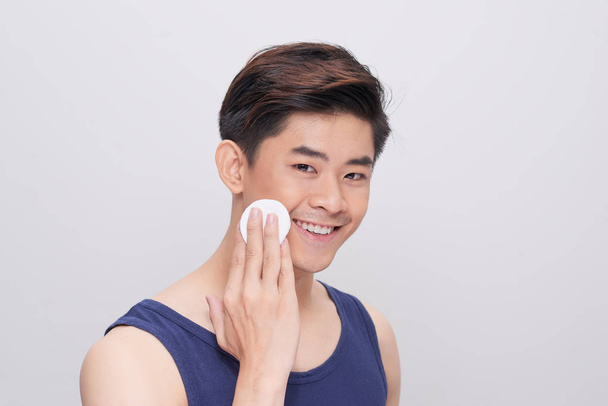 Handsome man cleaning face skin with batting cotton pads over gray background and looking at camera - Photo, Image