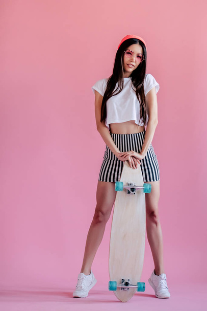 Young asian teenage girl on pink background. Stylish skater girl with skateboard isolated. - Photo, image