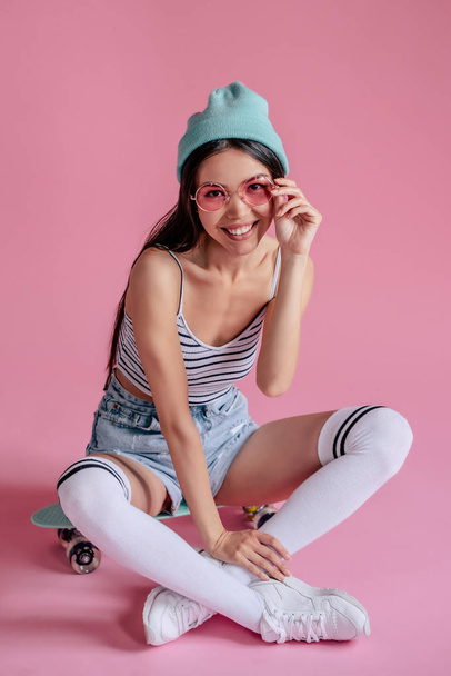 Young asian teenage girl on pink background with pink glasses. Skater girl in shorts with skateboard isolated. - 写真・画像