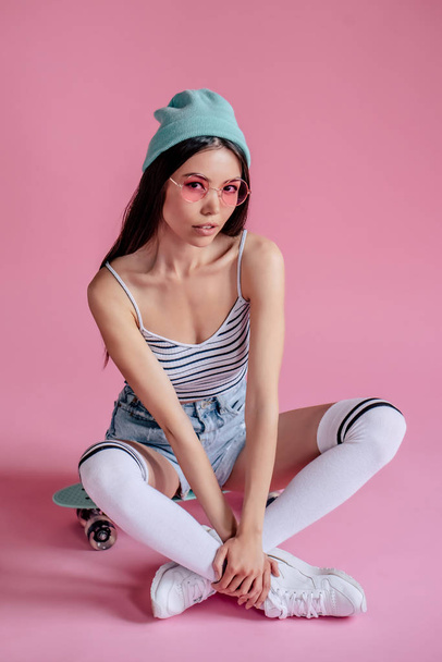 Young asian teenage girl on pink background with pink glasses. Skater girl in shorts with skateboard isolated. - Foto, Imagen