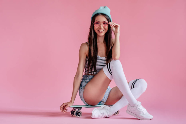 Young asian teenage girl on pink background with pink glasses. Skater girl in shorts with skateboard isolated. - Photo, image