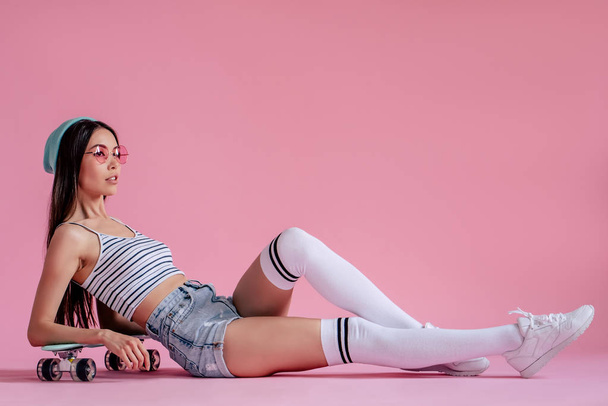 Young asian teenage girl on pink background with pink glasses. Skater girl in shorts with skateboard isolated. - Foto, afbeelding