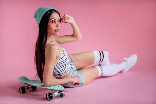 Jeune adolescente asiatique sur fond rose avec des lunettes roses. Skater fille en short avec planche à roulettes isolé
. - Photo, image