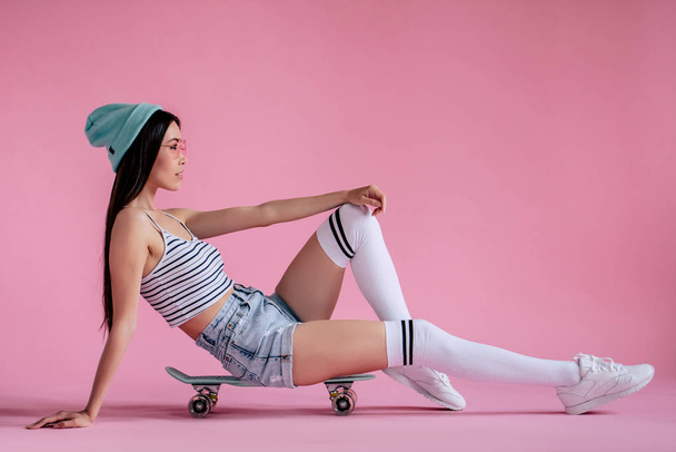 Young asian teenage girl on pink background with pink glasses. Skater girl in shorts with skateboard isolated. - Photo, Image