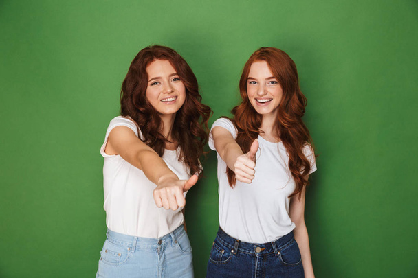 Portrait of two young redhead women 20s in casual wear smiling at camera and showing thumbs up isolated over green background - 写真・画像