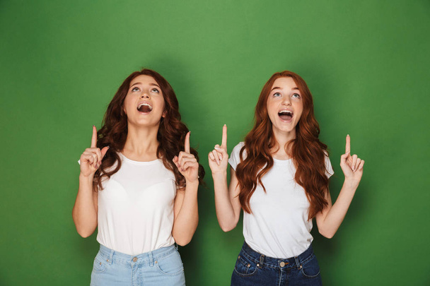 Portrait of two pretty women with red hair in white t-shirts smiling and pointing fingers upward in excitement isolated over green background - Valokuva, kuva