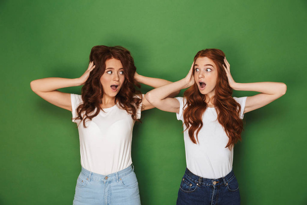 Portrait of two scared or uptight women with red hair in white t-shirts looking at each other and covering ears isolated over green background - 写真・画像