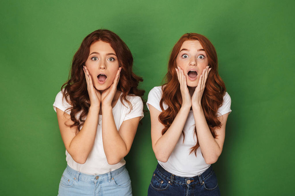 Photo of two caucasian redhead women 20s in white t-shirts touching cheeks with open mouth and bulging eyes isolated over green background - Foto, immagini