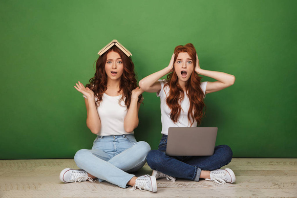Portrait of two shocked young redhead girls sitting on a floor with laptop computer and paper book isolated over green background - 写真・画像