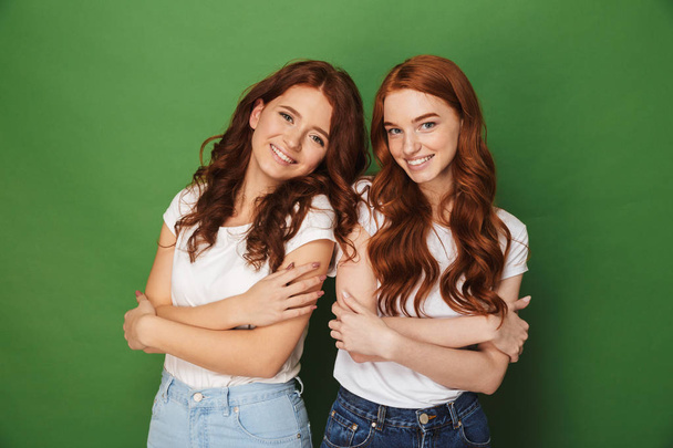Portrait of two adorable girls 20s with ginger hair in casual wear smiling and looking at camera with arms folded isolated over green background - Foto, immagini
