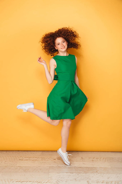 Full length portrait of a smiling young redhead woman in dress posing while standing isolated over yellow background - Foto, immagini