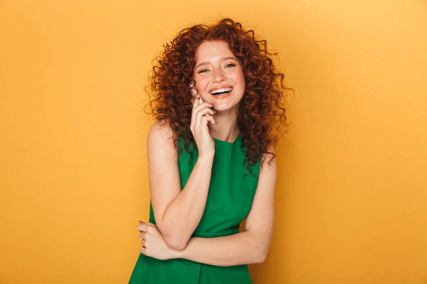 Portrait of a beautiful redhead woman in dress talking on mobile phone isolated over yellow background - Fotoğraf, Görsel