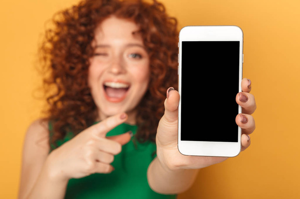 Portrait of a happy redhead woman in dress pointing finger at blank screen mobile phone isolated over yellow background - Valokuva, kuva