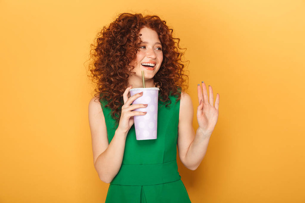 Portrait of a smiling redhead woman in dress drinking from a plastic cup a nd waving with hand isolated over yellow background - Photo, Image
