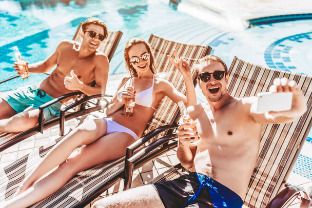 Group of young friends is lying on a chaise-longues near swimming pool and smiling while taking selfie on a smart phone. Having rest together and drinking beer. - Photo, Image