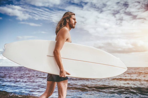 Young handsome man with long hair is walking on beach with white surfboard in hands. - Foto, imagen