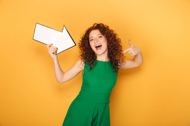Portrait of a cheerful redhead woman in dress pointing away with an arrow isolated over yellow background - Foto, afbeelding