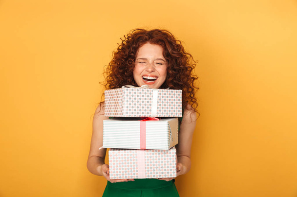 Portrait of a cheerful redhead woman in dress holding stack of present boxes isolated over yellow background - Foto, imagen
