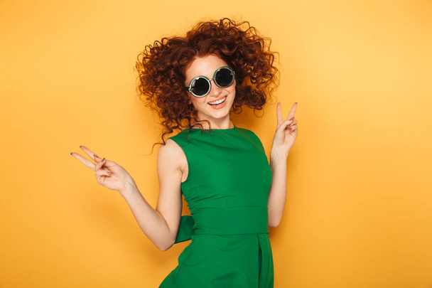 Portrait of an excited redhead woman in dress and sunglasses celebrating success isolated over yellow background - Valokuva, kuva